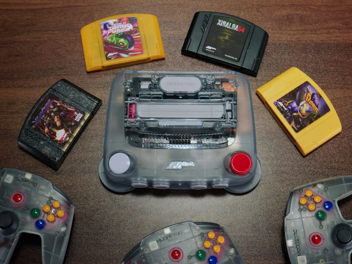 ModRetro M64 console in clear smoke plastic, with controllers and cartridges around it
